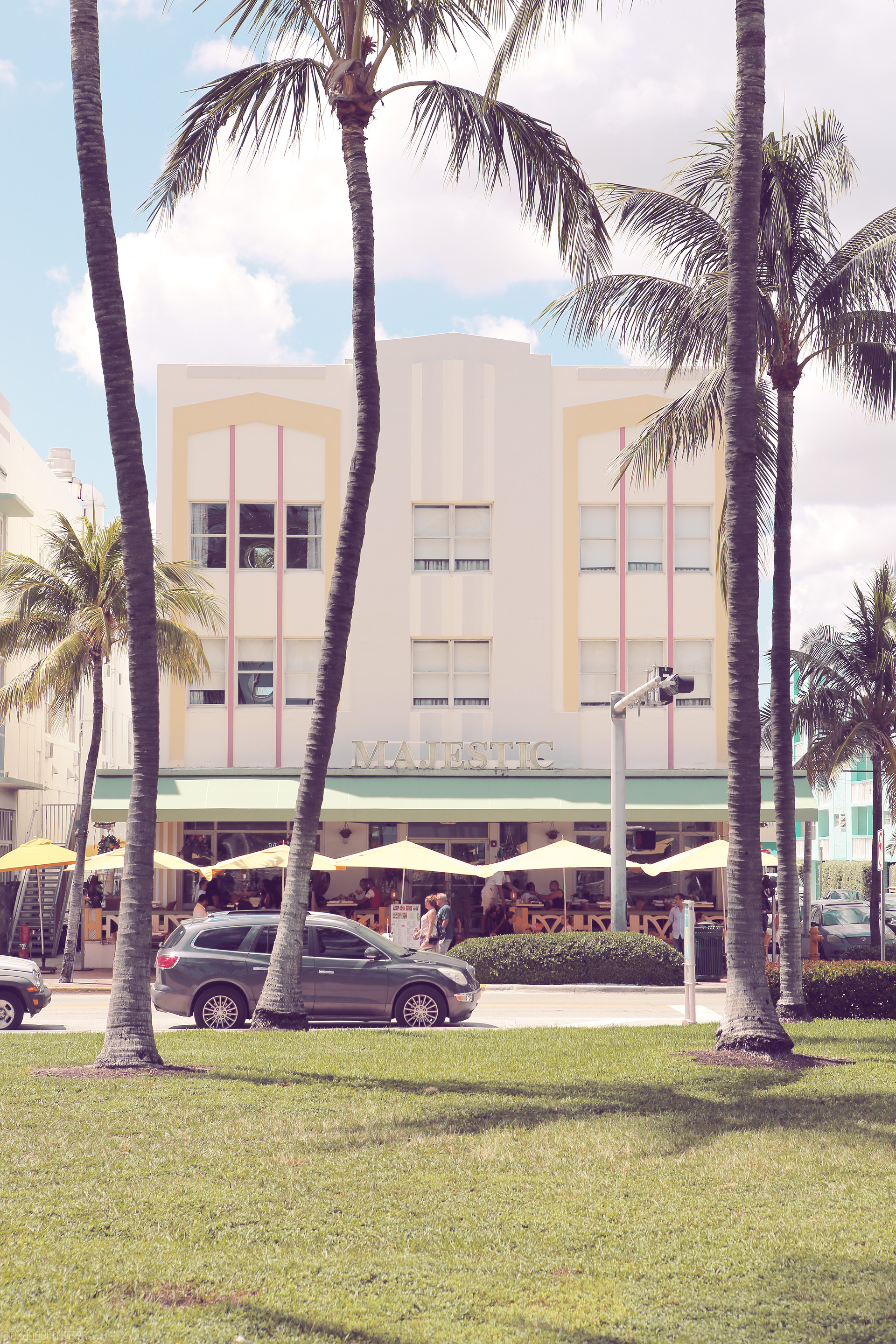 Tropical Facade Foto von A pastel Art Deco building on Ocean Drive, Miami, framed by palm trees under a sunny sky.