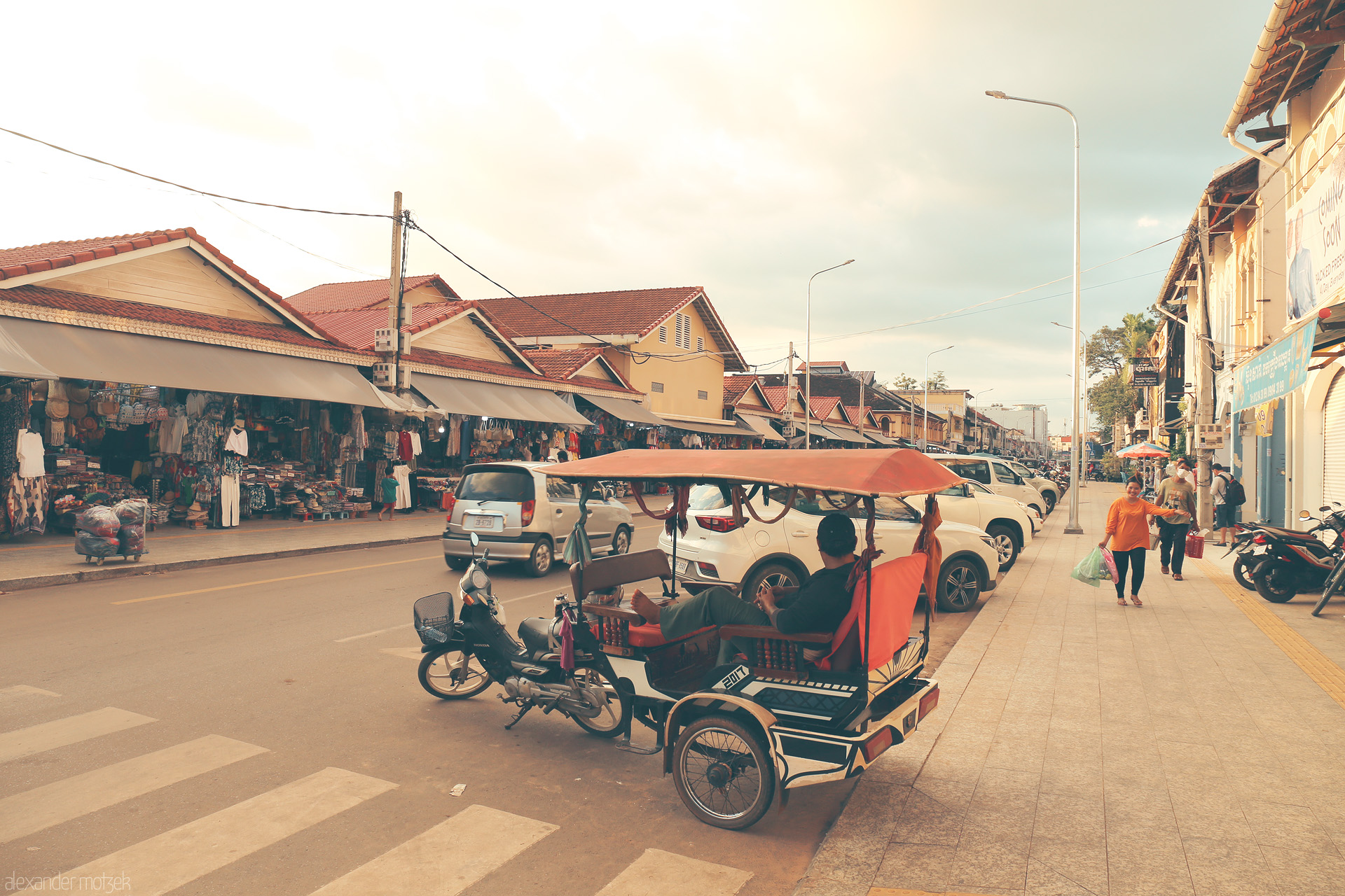 Foto von A tuk-tuk driver lounges by Siem Reap’s bustling Psar market, warm sunlight bathing local shops and vibrant street life.