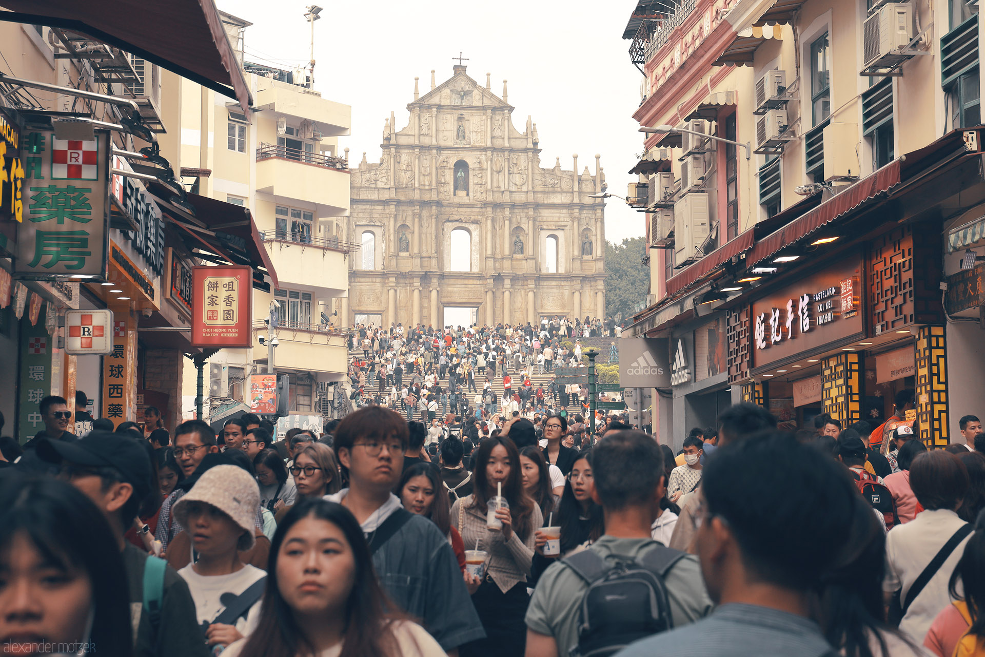 Foto von Crowds flow up Rua de Sao Paulo to the Ruins of St. Paul’s, Santo António, Macau, framed by shop signs and pastel balconies.