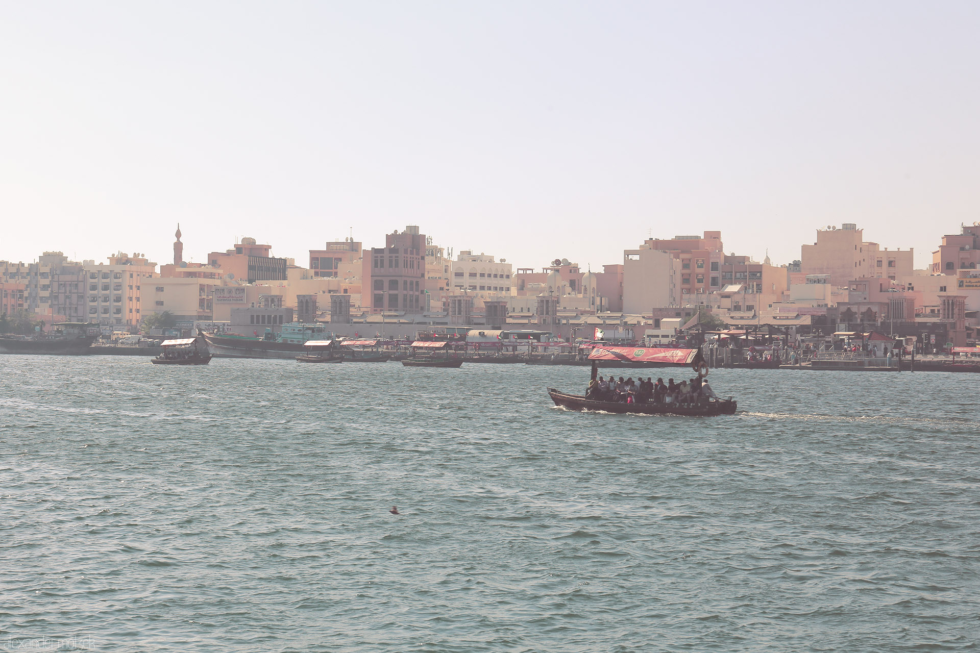 Abra Dreams, Deira Foto von Traditional abra ferries glide across Dubai Creek, framed by old Deira’s pastel skyline and the subtle haze of an Emirati afternoon.