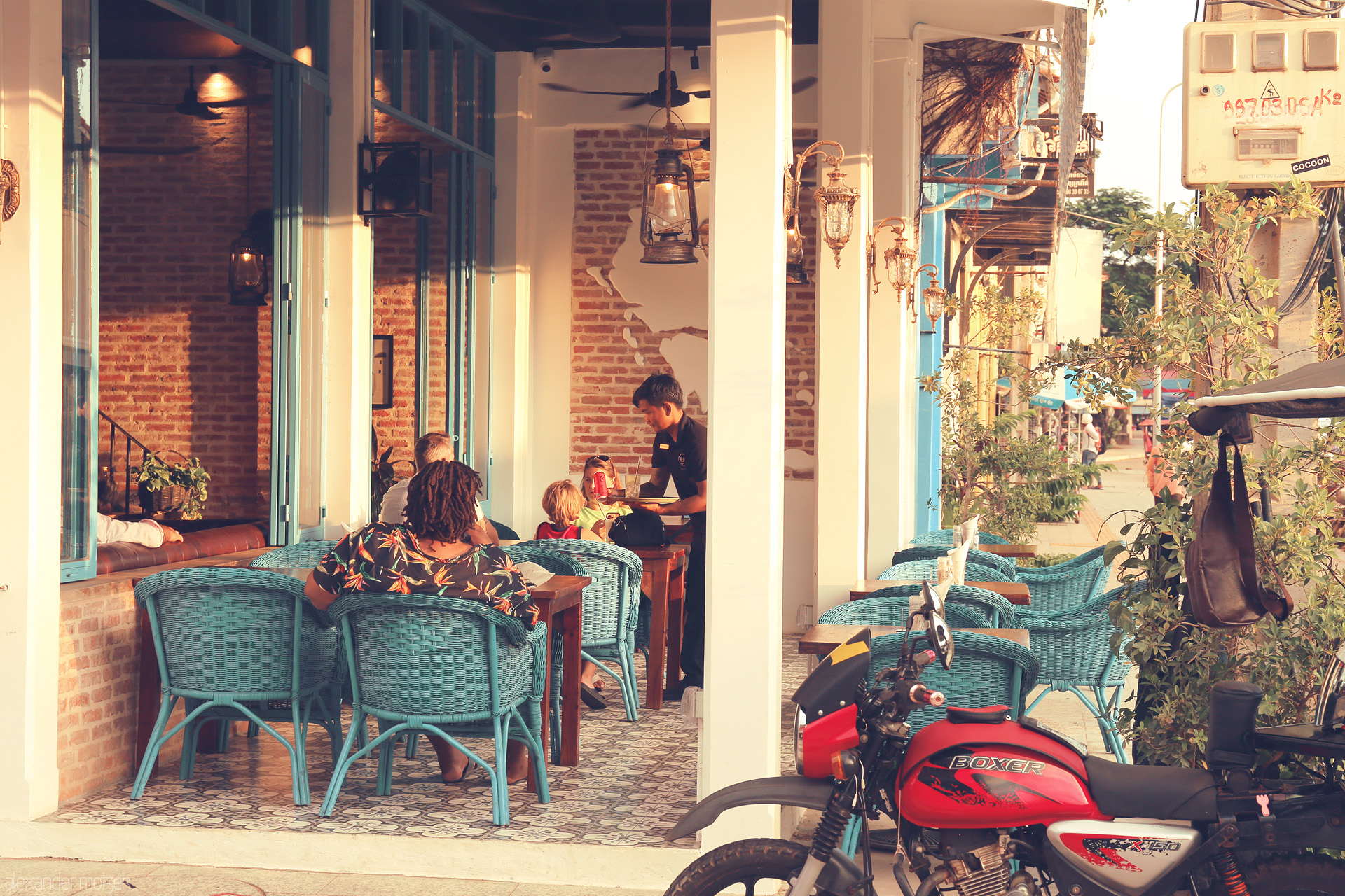 Foto von Evening light spills onto a cozy Siem Reap café terrace, turquoise chairs, laughter, and a red motorbike capturing Khmer street charm.