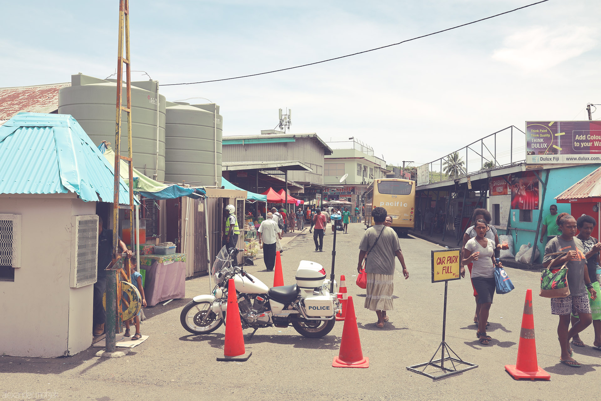 Foto von Midday bustle on Sigatoka’s market street—stalls, shoppers, a Pacific bus and a parked police motorbike amid bright cones, Fiji.