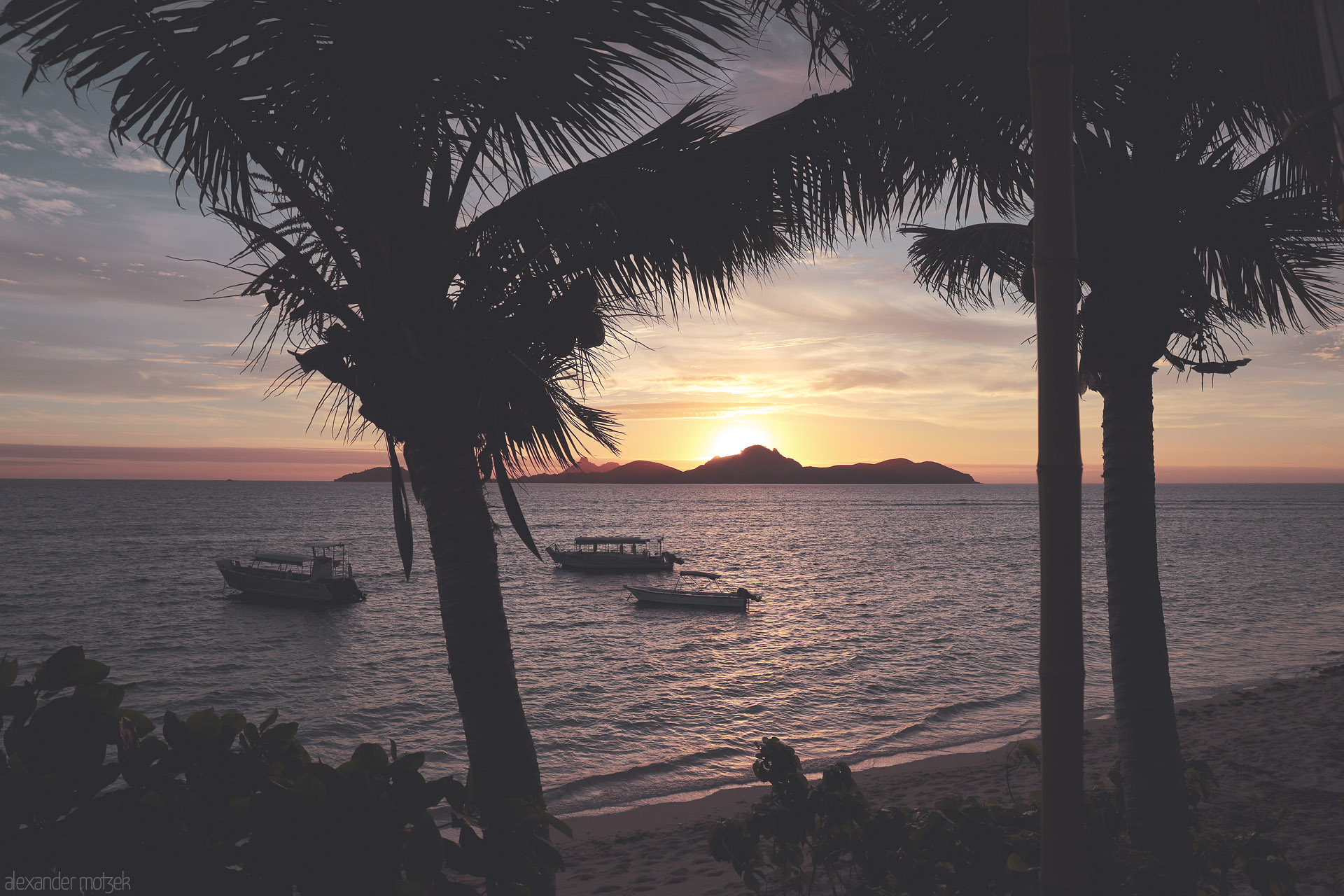 Foto von Palm silhouettes frame a Tokoriki sunset over the Mamanuca Sea, Fiji, with anchored boats rocking on gentle waves and a dark island horizon.