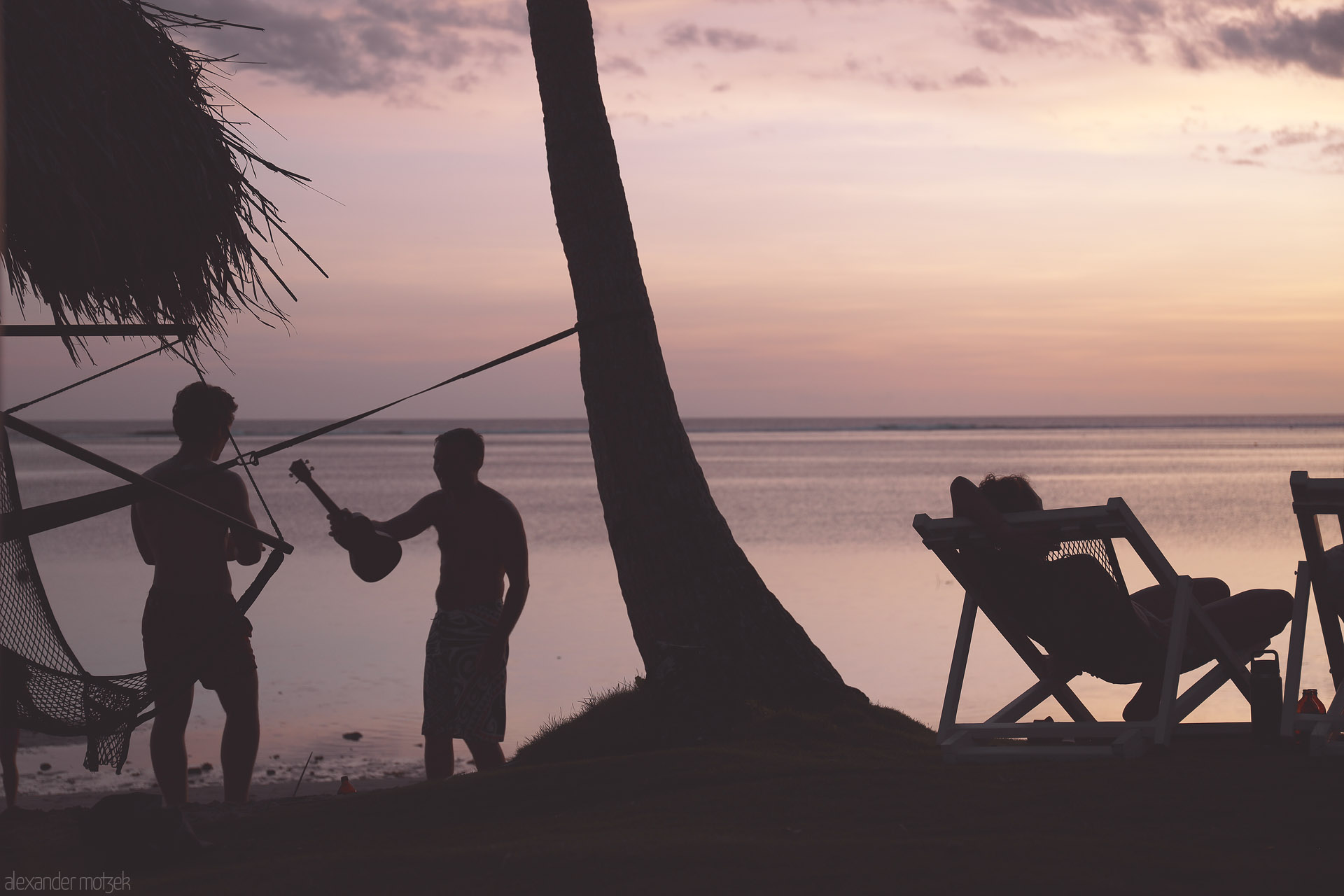 Foto von Silhouettes trade a ukulele beside a hammock and palm as a traveler reclines, watching Komave, Fiji's lagoon glow at sunset.