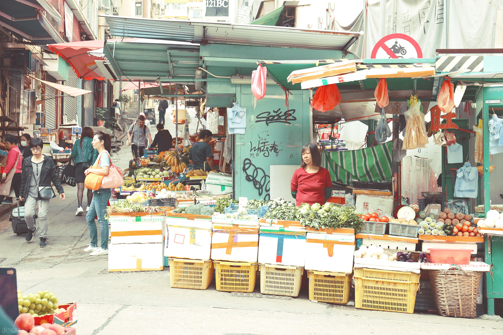 Foto von Open-air produce stall on Hong Kong Island—bananas, greens and tomatoes stacked high as shoppers weave through a narrow market lane.