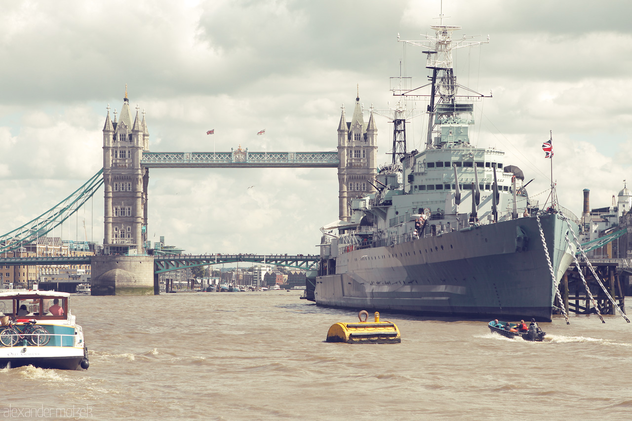 Thames Sentinel Foto von Iconic Tower Bridge and a majestic warship anchored along the Thames in London. A blend of history and modernity under cloudy skies.