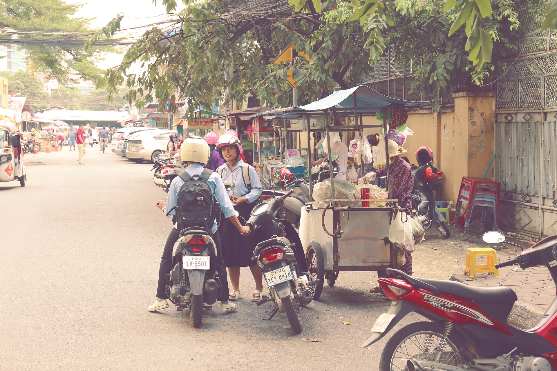 Foto von Local life unfolds on a Phnom Penh street: scooters, school uniforms, and a bustling food cart beneath lush trees in Cambodia's capital.