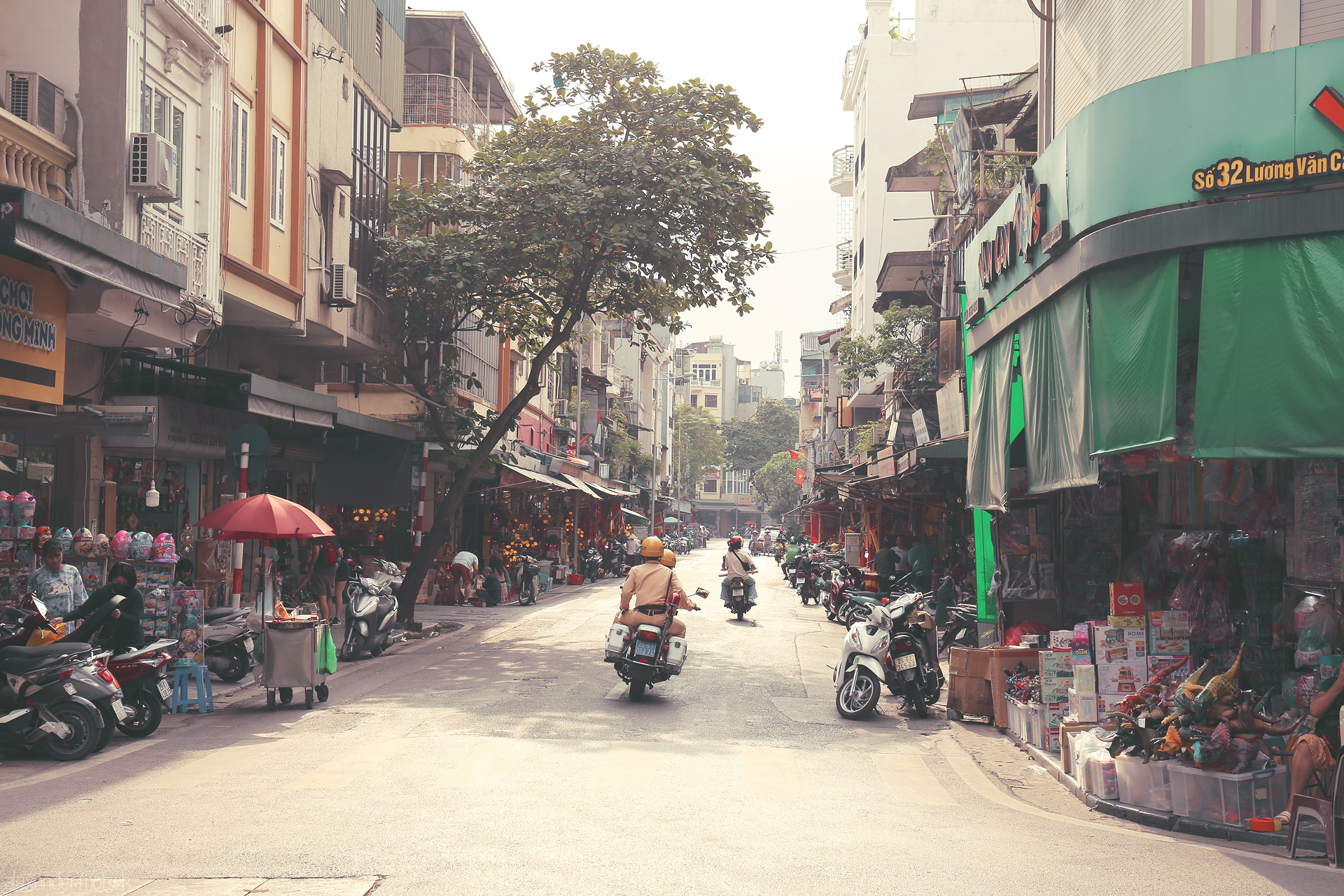 Foto von Morning scooters glide through sun-drenched streets of Phuong Hàng Dào, Hanoi, where market life hums beneath leafy branches.