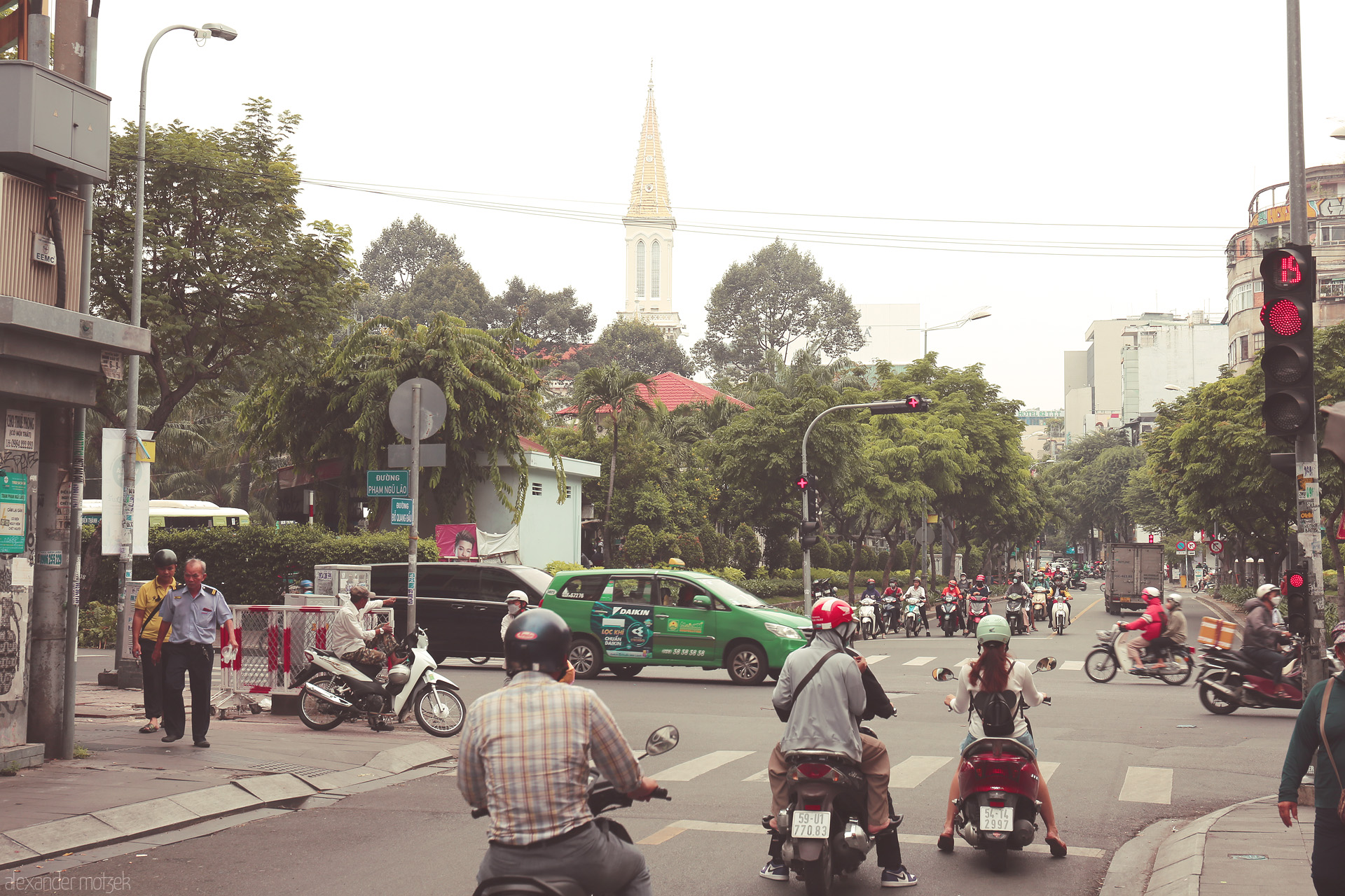 Foto von Motorbikes pause at a red light on Pham Ngu Lão, Saigon, as a church spire rises above lush trees in Ho Chi Minh City.
