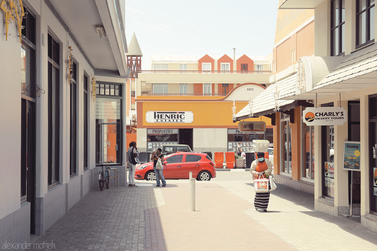 Foto von Urban street in Swakopmund under a clear blue sky, showing architecture, a bright red car, and local passerby.