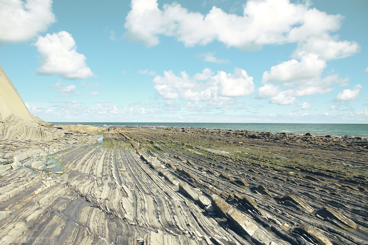 Foto von Layered rock formations of Flysch lead to the sea in Deba, Basque Country, under a vast, cloud-strewn sky.