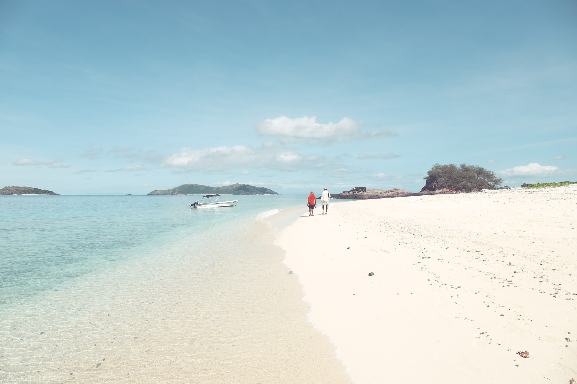 Foto von Two travelers trace Monoriki’s white-sand curve, Fiji; glassy lagoon and a lone boat—Cast Away was filmed here.